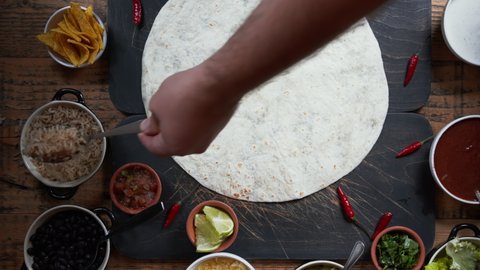 Top down view of making a burrito. Adding meat, salsa, herbs and fresh vegetables to tortilla and making traditional Mexican cuisine dish. Overhead view of food preparation