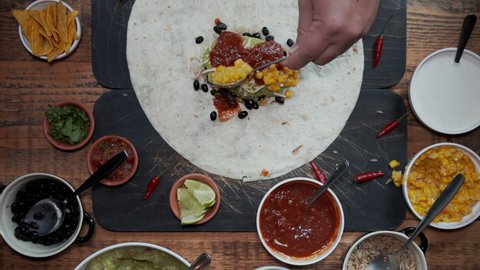 Overhead view of adding ingredients to tortilla and making a burrito. Top down view of adding fresh vegetables, sauces and herbs into tortilla and wrapping it up. Cutting burrito in half