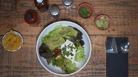 Overhead of traditional Mexican salad in the bowl placed on served wooden table. Fresh vegetables and salsa in a bowl placed next to a bowl of tortilla chips, limes and parsley