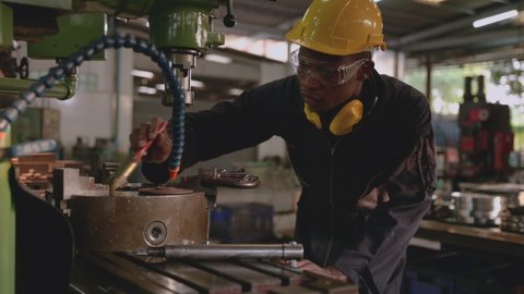Professional technician african american man in uniform wearing hardhat and goggles with earmuff is standing cleaning and protect rust by oil coating to chucks of milling machine in factory industrial