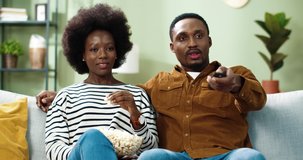 Young happy african american couple is sitting at home on couch and watching movie on TV and eating popcorn. Man uses remote control to switch channels.