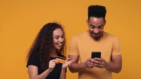 Smiling young couple friends african american man woman 20s isolated on yellow background studio. People lifestyle concept. Using mobile cell phones typing hold credit bank card doing winner gesture