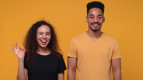 Cheerful smiling young couple two friends african american man woman 20s in basic t-shirts posing isolated on yellow color background studio. People lifestyle concept. Showing ok okay gesture blinking