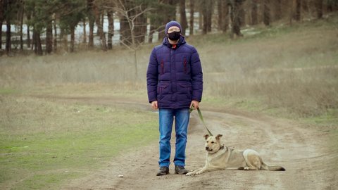 Man in black protective face mask for coronavirus prevention and glasses stand outdoors with dog on leash in near forest in winter during pandemic. Stop dangerous COVID-19 infection