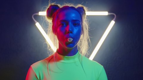 Girl inflates bubble of gum. Young woman looking at the camera in the neon light of the club.  Beautiful futuristic lady close-up. Camera zoom. 