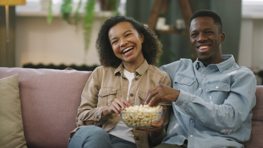 Slow-motion medium shot of young cheerful afro couple laughing while watching film on TV, sitting together on couch at home eating popcorn Royalty-Free Stock Footage #1067603420