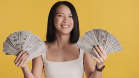 Beautiful Asian woman looking confident showing wide of money on camera and rejoicing isolated on yellow background. Successful concept