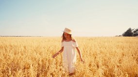 joyful girl in white dress and straw hat walking in field and touching straw hat