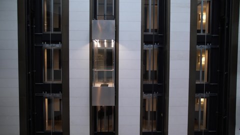 Close-up of modern transparent passenger elevator shafts in luxury hotel. Busy on cellphone respectable businessman with travel bag entering lift cabin, starting it with guest card and going down