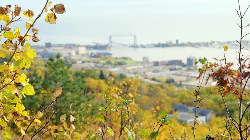 Focus shifts from nearby leaves to the Duluth Minnesota harbor and aerial lift bridge and the Great Lake Superior, in landscape clip on a mostly cloudy day in autumn.