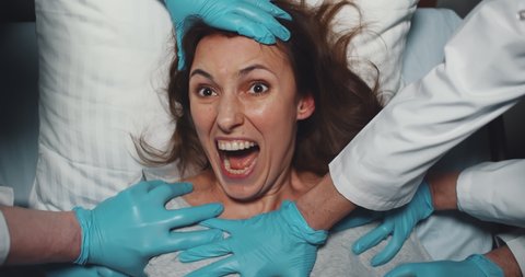 Top view of doctors holding female psycho patient on bed in hospital. Medical workers in latex gloves holding screaming insane woman trying to escape