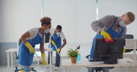 Team of diverse young cleaning service professionals in safety mask at work in office. Multiethnic janitors wearing protective mask and uniform cleaning modern office