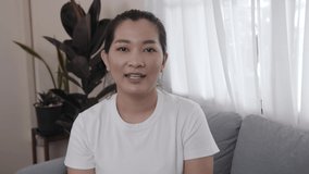 Happy Asian young female sitting on sofa talking to camera for communication to viewers shooting vlog and video, smiling ethnic girl greeting talking having a conversation or video call at home