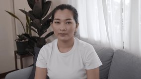 Happy Asian young female sitting on sofa waving to camera say hello to viewers shooting vlog or video, smiling ethnic girl greeting talking having a conversation or video call at the laptop