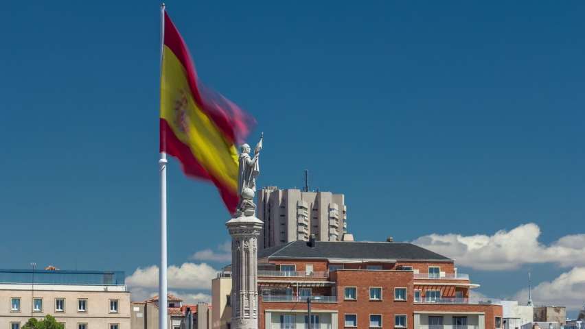 Spanish flag waves behind statue of Christopher Columbus timelapse with clouds on blue sky, plaza de Colon in Madrid, Spain
