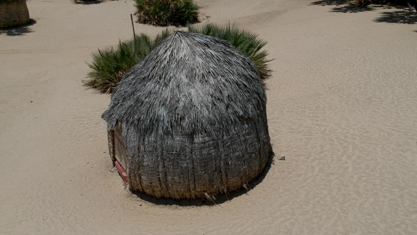 Round traditional hut made from Palm tree leaves of the Turkana people in the middle of small group of huts on sandy landscape camera moving away from hut Aerial.