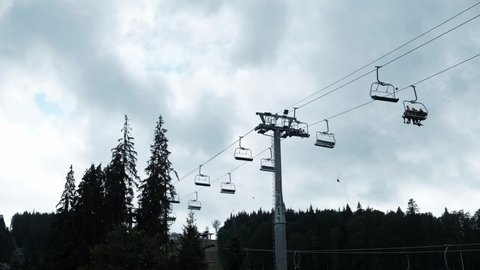 View from the ground at a ski lift in the summer. Overcast gray cloudy sky, dark moody weather, electrical cars with tourists flying up and down the rope into the forest and mountains. Weekend outdoor