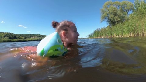 A happy little girl is floating on the river in inflatable armbands, on a bright sunny day, against a background of blue sky and green reeds. Close-up. View from the water surface. 4K
