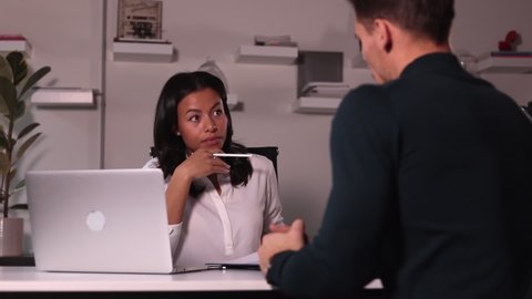 Woman talking to man during hiring interview at table in modern office spbi. Young female specialist has dialogue with male candidate and looks at resume, sitting at desk with laptop in light interior