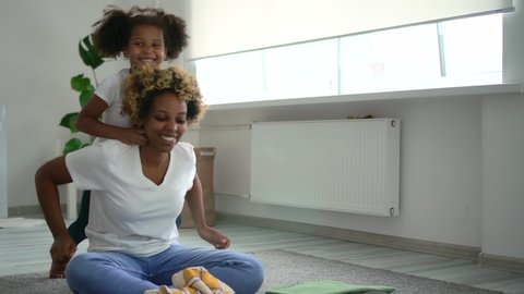 Young mother and cute daughter have fun and sit on floor in laundry room. American African woman and little girl hugging and talking with happy smiles while sitting in light interior. Cheerful: film stockowy