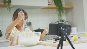 Asian girl mixing ingredients to make cupcakes in kitchen at home whilst vlogging on mobile phone - shot in slow motion