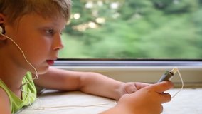 Closeup view profile video portrait of happy little white boy travelling by train and using mobile phone while sitting at table in moving wagon of train. Green summer trees passing by in background