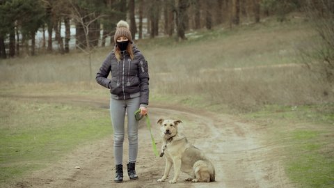 Woman in black protective face mask for coronavirus prevention stand outdoors with dog on leash in near forest in winter. Stop dangerous COVID-19 infection