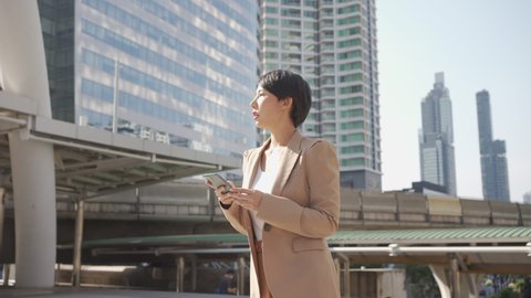 Asian Short hair businesswoman using smartphone, walking on street middle of building in capital city. Confident woman in suit walking and looking on the phone with cheerful smile. Low angle view. Video de stock