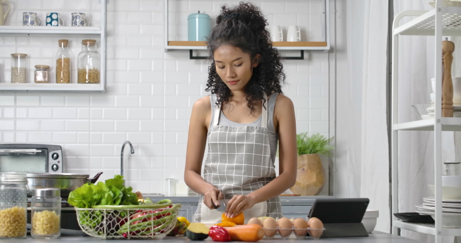 A young, happy and beautiful Asian woman with curly long hair wearing grey gingham apron. She slide vegetable on chopping block with knife and looking at the tablet for food recipe in modern kitchen. Royalty-Free Stock Footage #1067656019
