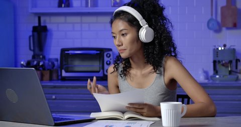 A young, exhausted and tried Asian woman sitting in modern kitchen, She's wearing headphone and using computer for making a video call with her co-worker. Working late at night and New normal concept. Video stock