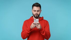 Smiling pensive young bearded man 20s in casual red streetwear hoodie isolated on blue background studio. People lifestyle concept. Using mobile cell phone typing sms message browsing looking aside up