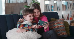 Lovely young couple of excited children hugging on couch using mobile phone watching videovlog videos having fun at home. Entertainment. Families.
