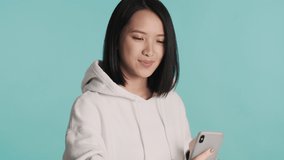 Pretty Asian girl dressed in white hoodie looking cute taking selfie on smartphone over blue background. Modern technology concept