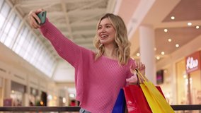 Blogger influencer speaking to camera phone recording vlog for channel. Young woman talking in video conference by smartphone holding colored gift bags. Smiling girl using smartphone in mall.