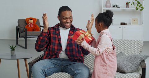 Cute faro kid daughter make surprise present for dad receive gift box sit on sofa, black man father covering closing eyes while little child girl congratulating parent on fathers day birthday at home