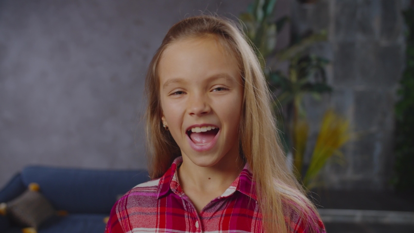 Headshot of lovely hilarious elementary age girl in casual clothes laughing infectiously, rejoicing and expressing carefree mood, fun, happiness, joy while posing in home interior.