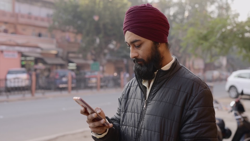 Static shot of a young Indian office worker man wearing a turban on the head with a long beard using a mobile phone to type a text message standing by the corner of a street during morning hours