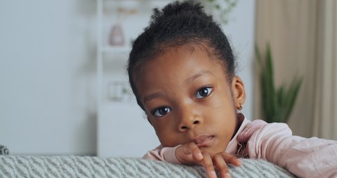 Portrait of afro american child little girl with dark skin and large wide open eyes looks at camera emotions of sadness stress, close-up of calm face of toddler indoors, orphan kid missing his family