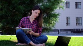 Attractive girl works for a laptop sitting on the grass in a city park on a summer morning, shooting outdoors in an urban area. Happy sunny summer day in the park and video calling. Online learning