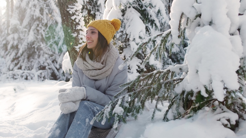 Portrait of a beautiful female in a winter forest, cheerful young woman sitting next to a tree and enjoying the snowfall during a winter walk in nature, 4k slow motion.