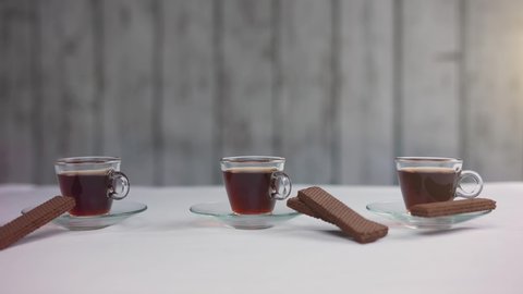 Dolly shot of three glass cups of espresso coffee or tea with chocolate wafer biscuits on a white table and background. Cafe, coffee shop, kitchen. 