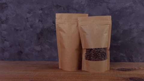 Hand in protective gloves holds brown paper pouch bags mockup with coffee beans wooden background 4K. Packaging for food delivery goods template. Kraft pack with clasp window tea leaves weight product