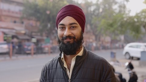 Static shot of a young Indian office worker man wearing a traditional turban on the head with a long beard looking at the camera, standing by the corner of a street during morning hours