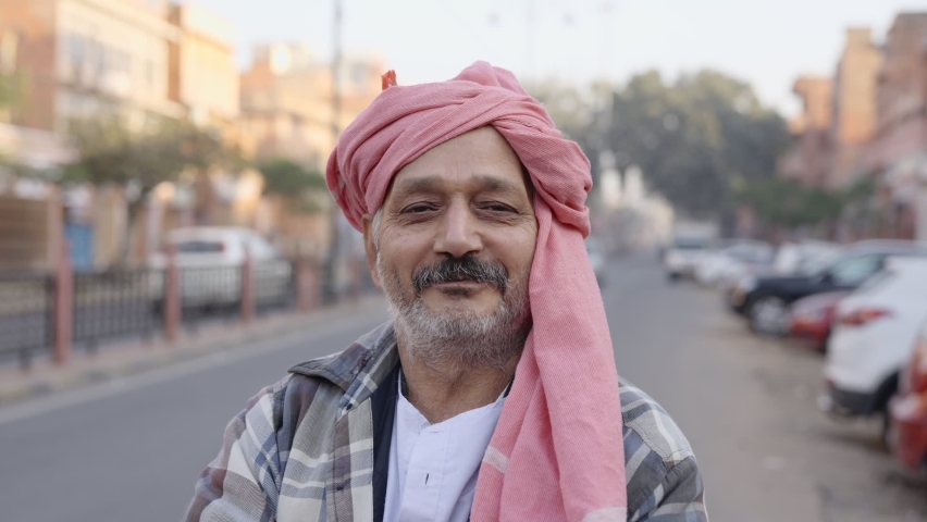Close view static shot of an old aged Indian Asian traditional common man wearing a turban on his head standing at the side of an urban local street and looking at the camera 