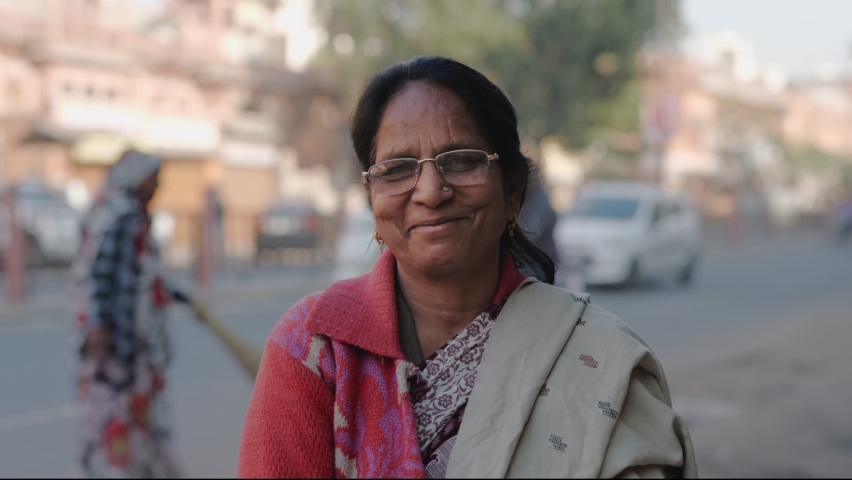 Close static shot of a middle aged Indian Asian common woman wearing specs and a traditional saree with a smile on the face standing at the side of an urban local street and looking at the camera  | Shutterstock HD Video #1067701352