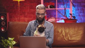 Young hipster man sitting on couch with cute pug dog having video call on laptop. Bearded guy in headphones waving hand at camera talking on video conference and holding pet