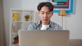 Young asian man working remotely on laptop at home office. Portrait of concentrated freelancer typing on computer sitting at home. Korean student guy e-learning in living room