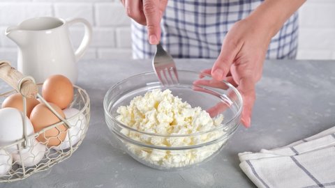 Cottage cheese or farmer's cheese in a glass bowl on the kitchen table. Woman hand stirring crumbly fresh white cheese