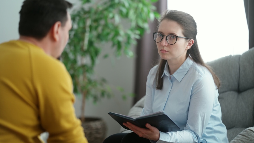 The young man is sitting in the psychologist's office, telling her about his problems. A beautiful woman psychologist consulting her male patient writes notes in a notebook | Shutterstock HD Video #1067705378
