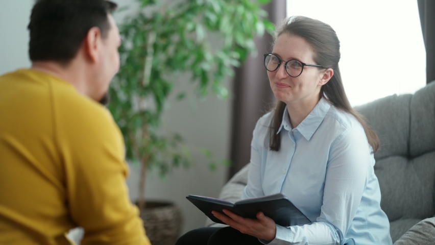 The young man is sitting in the psychologist's office, telling her about his problems. A beautiful woman psychologist consulting her male patient writes notes in a notebook | Shutterstock HD Video #1067705378